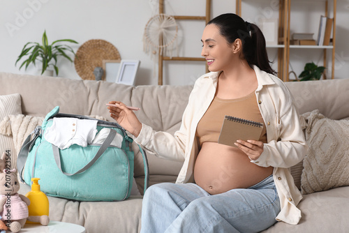 Young pregnant woman with checklist and maternity hospital bag sitting on sof...