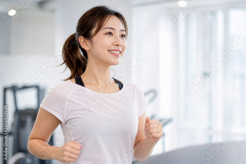 スポーツウェアを着用しフィットネスジムでランニングマシンを使って有酸素運動する笑顔の若い日本人女性、ダイエット・健康管理・運動習慣のイメージ