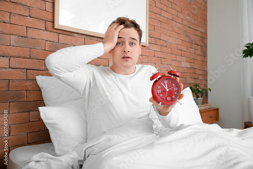 Shocked young man with alarm clock sitting in bed at home