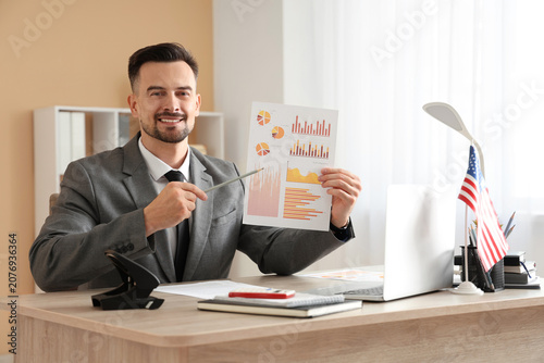 Handsome young happy businessman with laptop, diagrams and flag of USA in office