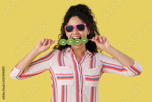 Beautiful young happy African-American woman holding stick with kiwi on yello...