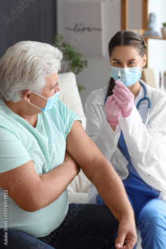 Nurse with injection and senior man in medical masks at home