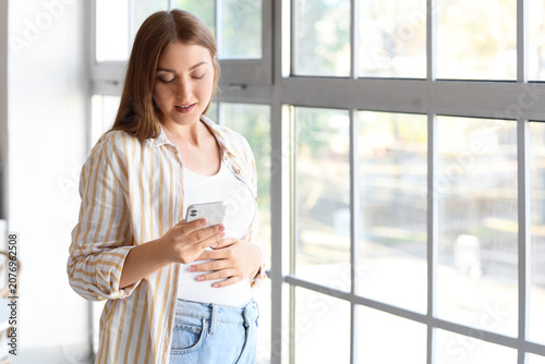 Young pregnant woman using mobile phone near window at home