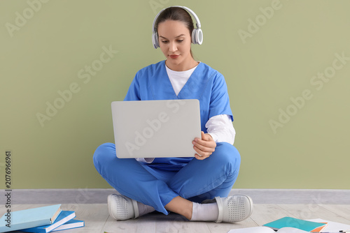 Female medical intern in headphones studying with laptop near green wall