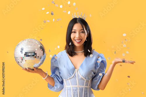 Happy fashionable young Asian woman with disco ball and confetti on yellow ba...
