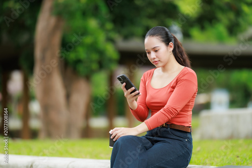 Young Asian woman is sitting outdoors in a park, holding a smartphone and a coffee cup, focusing on her device for communication, social media, or remote work activities