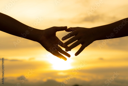Close up of two hands reaching out to hold each other against sunset sky background.
