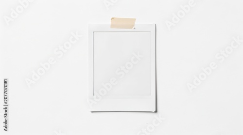 A blank instant photo with a white frame is taped to a white wall using beige masking tape.