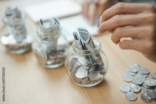 Fotografie Hand placing coins into savings jars with cash, representing savings goals, budgeting milestones, emergency funds, and personal financial planning for long-term stability and future success