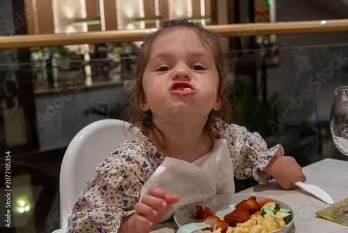 Schilderij op canvas A funny and cheeky little girl sits in a white high chair at a restaurant table, making a silly grimace with her mouth full of food