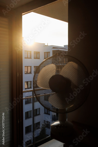 Photography An electric fan standing inside a room at sunrise on a hot summer morning, circulating cool air to improve ventilation during an extreme heatwave