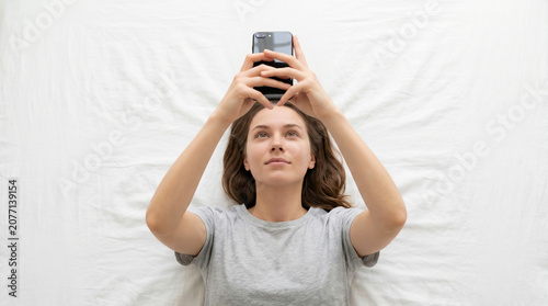 Young woman lying on white bed holding smartphone above her head, relaxed top view lifestyle shot with copy space for technology and bedtime themes