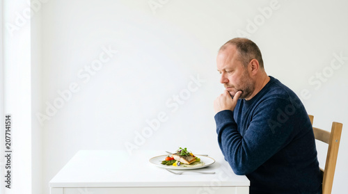 Pensive middle-aged man sitting alone at a white table looking at a small gourmet fish dish. Bright minimalist background with copy space. Perfect for diet, loneliness, or lifestyle concepts.