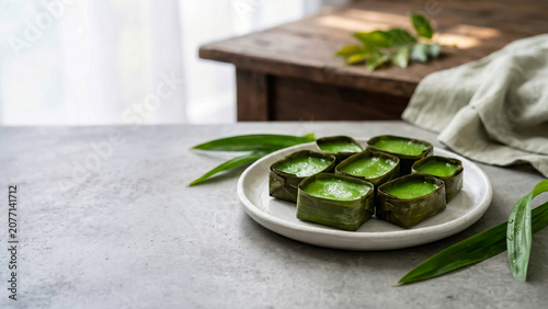 Traditional Asian green pandan dessert in banana leaf cups on a plate. Grey concrete table background with copy space. Ideal for Indonesian culinary, recipe blogs, and authentic food photography.