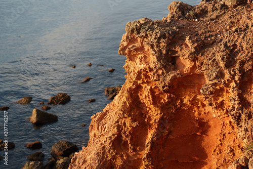 Close-up of a textured orange sandstone cliff face glowing in warm sunlight above the calm blue sea. This rugged coastal rock formation was photographed along the Portuguese shoreline.