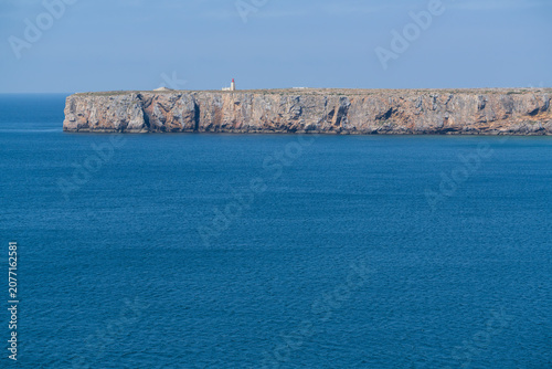 Dramatic limestone cliffs rise above the deep blue Atlantic Ocean along the rugged Algarve coastline in southern Portugal. A small lighthouse stands atop the distant headland under a clear sky