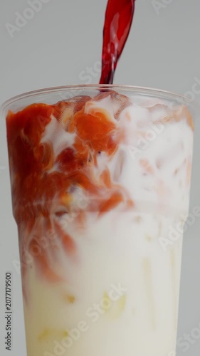 4K Close-up of dark red syrup or tea being poured into plastic cup filled with milk and ice creating a swirling layer effect isolated on light neutral background for drink commercial.