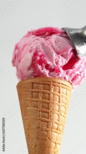 4K Close-up of metal scoop releasing perfect ball of vibrant pink strawberry ice cream onto crispy waffle cone, isolated on light gray background.