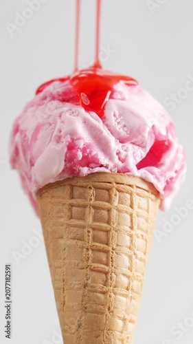 4K Close-up of strawberry syrup pouring over a scoop of pink ice cream on a waffle cone delicious dessert texture isolated on light background.