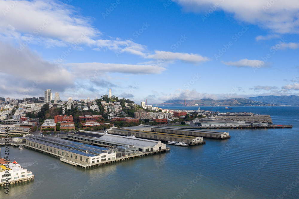 Obraz premium Panoramic aerial view capturing San Francisco's historic Embarcadero waterfront with piers, city architecture, Coit Tower, and the Golden Gate Bridge in the distance under a blue sky