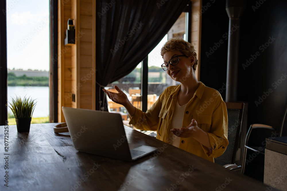 Fototapeta premium A Caucasian woman with short blonde hair and glasses is actively engaged in a video call on her laptop, gesturing with her hands while sitting at a wooden table