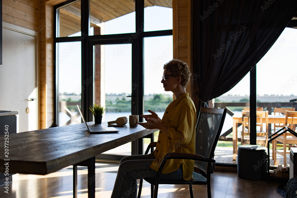 Fototapeta premium A Caucasian woman sits at a wooden table, engaged in a video call on her laptop with a scenic view of a lake and greenery in the background