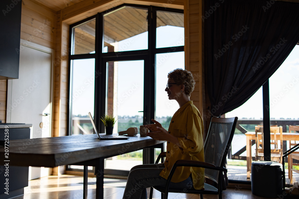 Fototapeta premium A Caucasian woman wearing glasses and a yellow shirt is sitting at a wooden table in a modern log cabin, engaged in a video call on her laptop