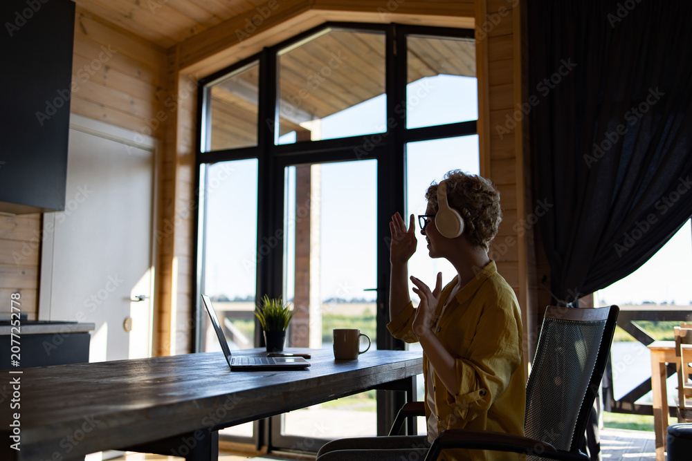 Fototapeta premium A Caucasian woman wearing headphones and glasses is engaged in a video call on her laptop while sitting at a wooden table in a room with large windows
