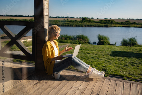 A Caucasian woman with short blonde hair wearing glasses and a yellow shirt is sitting on a wooden porch with a laptop and a mug of coffee