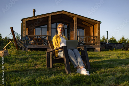 A young woman with short blonde hair and glasses sits in a chair outdoors with a laptop on her lap, enjoying the sunshine in front of a wooden cabin