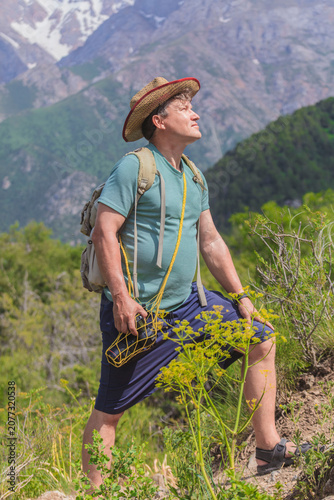 Man hikes mountain trail, eyes upward, smiling. Wears straw hat, backpack, and shorts. Surrounded by green shrubs, wildflowers, and trees. Mountains loom behind him, snow-capped peaks