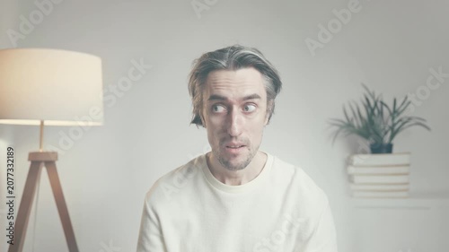Young man in white sweater sitting indoors in minimal room, looking at camera with varied expressions, illuminated by soft daylight and warm lamp