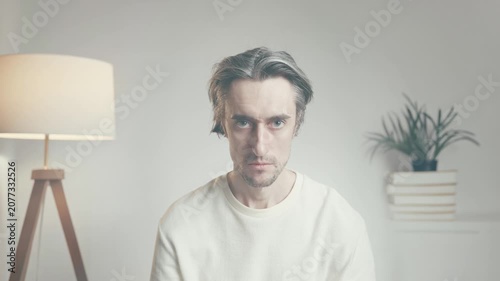 Young man with gray-streaked hair sitting indoors in white long-sleeve shirt, looking contemplative against minimalist backdrop with modern decor