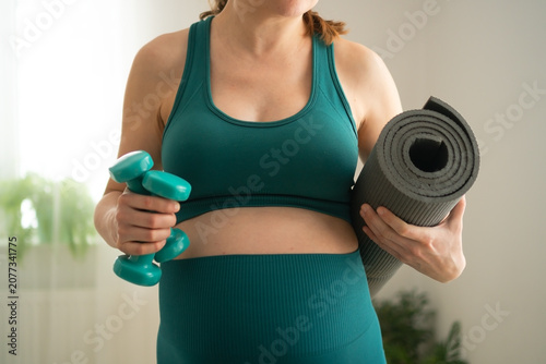 Pregnant woman holding dumbbell and yoga mat for prenatal fitness. Expecting mother shows discipline through consistent home workout. Mom-to-be stays focused maintaining regular exercise program