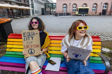 Smiling activists holding equality signs on a rainbow bench outside