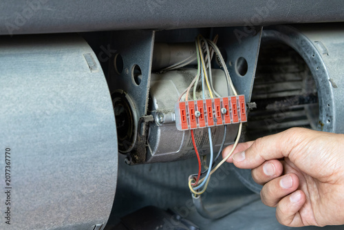 Technician hand checking electrical wiring and red terminal block inside opened air conditioning unit, between two cylindrical blower fans, HVAC maintenance.