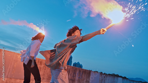 Fotografía Two adolescents stand on a rooftop, one holding a flaming torch, symbolizing youthful rebellion and the exuberance of adolescence against a sunset sky