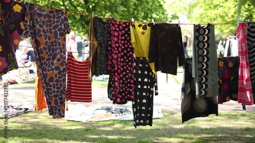 vibrant Marimekko dresses hanging on a clothesline at an outdoor flea market in a sunny Helsinki park, surrounded by lush green trees, blooming white blossoms, and shoppers.