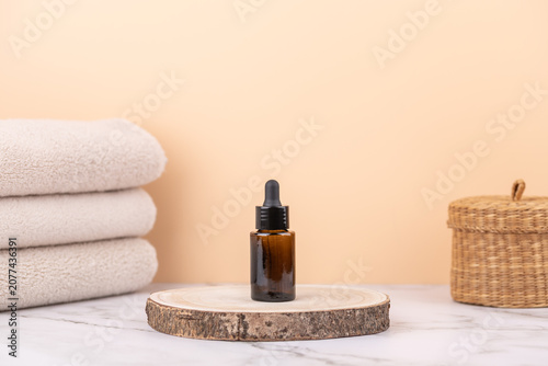Front view of amber glass dropper bottle on wooden base with beige towels and wicker basket on marble surface. Horizontal minimalistic composition with copy space. Concept of skincare and slow living