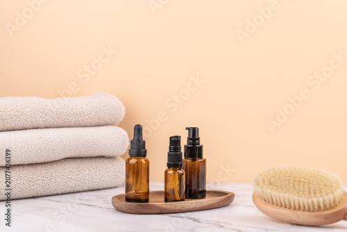 Three amber glass bottles with dispenser on a wooden tray, next to a massage brush and beige towels. Peach background and marble surface. Horizontal image with copy space
