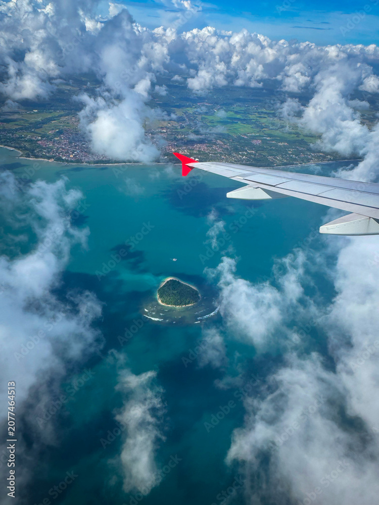 Naklejka premium AERIAL: Tip of an airplane wing above turquoise coastal water, drifting clouds and a small island. Traveling by plane over tropical coastline of island of Sumatra, Indonesia, with vivid sea colors.