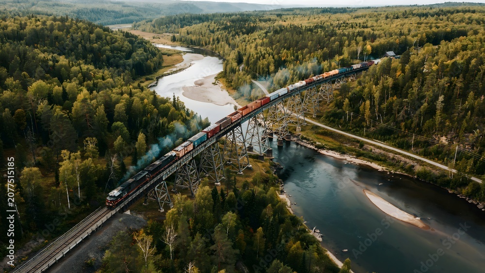 Fototapeta premium Aerial view of a colorful freight train crossing a lush green river valley with dense forest and rolling hills