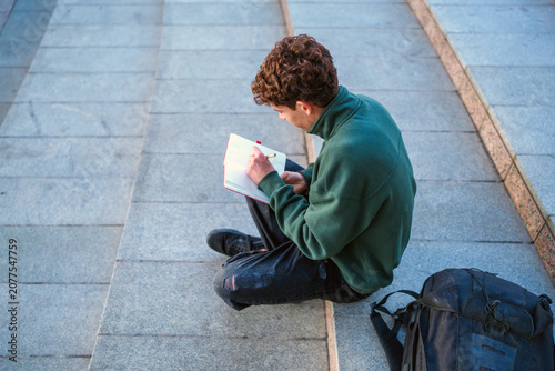 Young man sitting on outdoor steps, deeply engaged in writing notes in an open book with a pencil