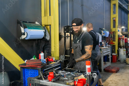 Tool cart stands near yellow beam inside repair workshop area. Bearded mechanic sorts sockets spraying cans as prepares parts for service task