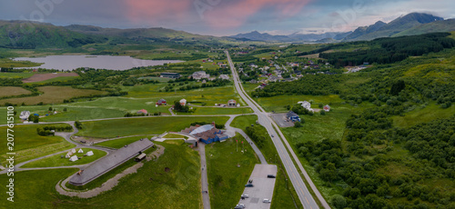 Photos Aerial view shows Lofotr Viking Museum longhouse, tents, modern building, and parking in Lofoten, Norway at dusk, with road to cloud brushed mountains and lake