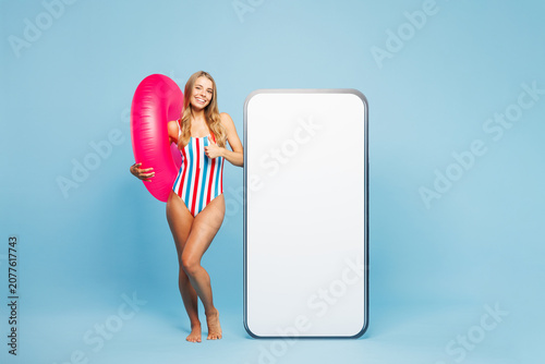 Full body young fun woman wear bikini swimsuit rest near hotel pool big huge blank screen area mobile cell phone show thumb up isolated on plain blue background. Summer vacation sea sun tan concept.
