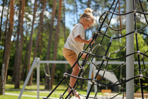 A young child ascends a rope climbing frame at a playground in the park. The sun shines down as trees provide background greenery