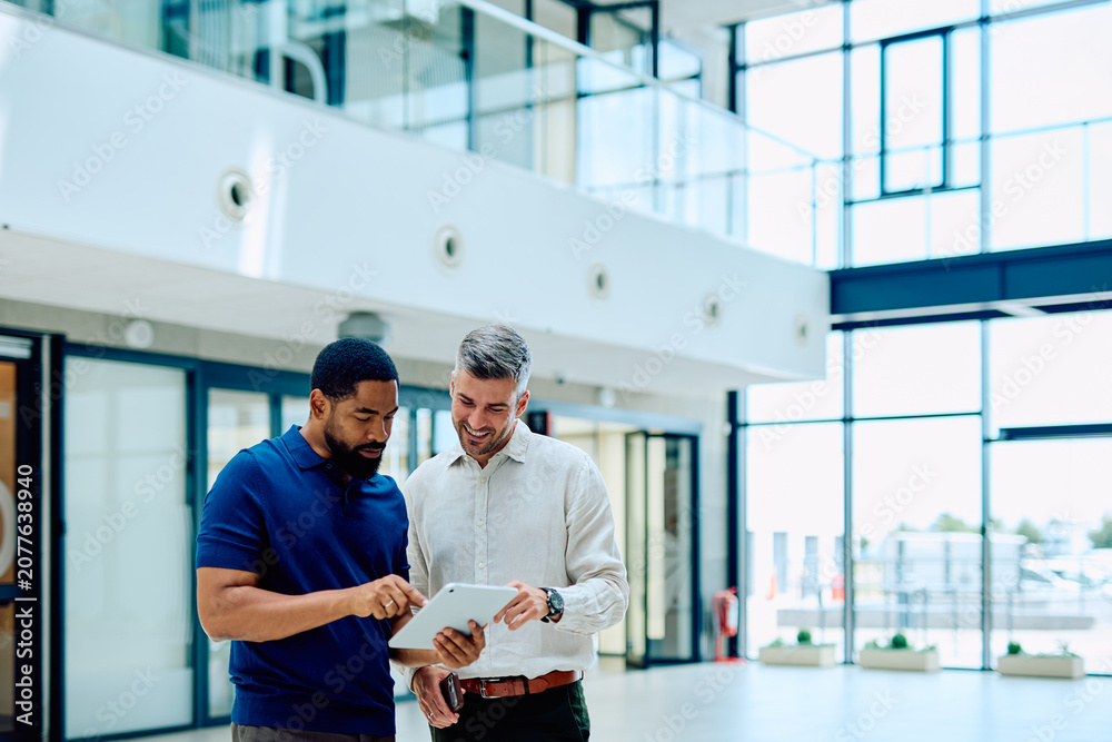 Fototapeta premium Two diverse businessmen collaborating on a project, analyzing data on a tablet in a contemporary office setting, sharing ideas