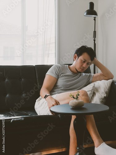 Young Thai man leaning forward tiredly on table from black sofa in relaxed apartment in a quiet minimalist Thai apartment with natural daylight