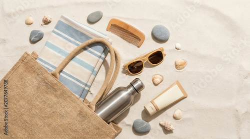 Minimal summer beach essentials arranged on soft sand, featuring tote bag, reusable bottle, sunglasses, towel, and skincare accessories in a clean travel flat lay.